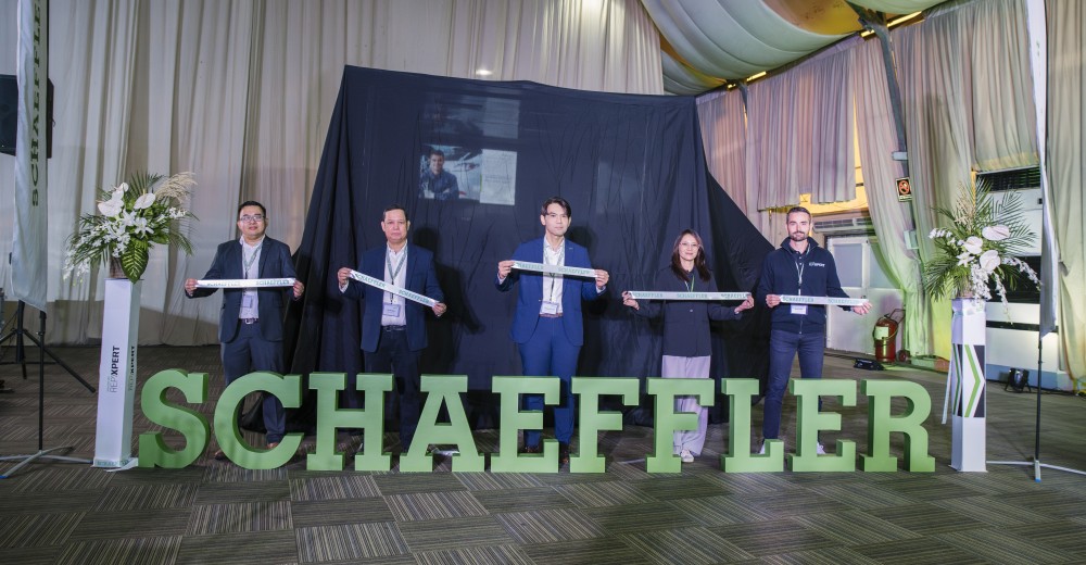 A group of five people is standing indoors in front of a large sign displaying the word "SCHAEFFLER" in green block letters. They are each holding a white ribbon with the Schaeffler logo on it, standing side by side in a ribbon-cutting ceremony.